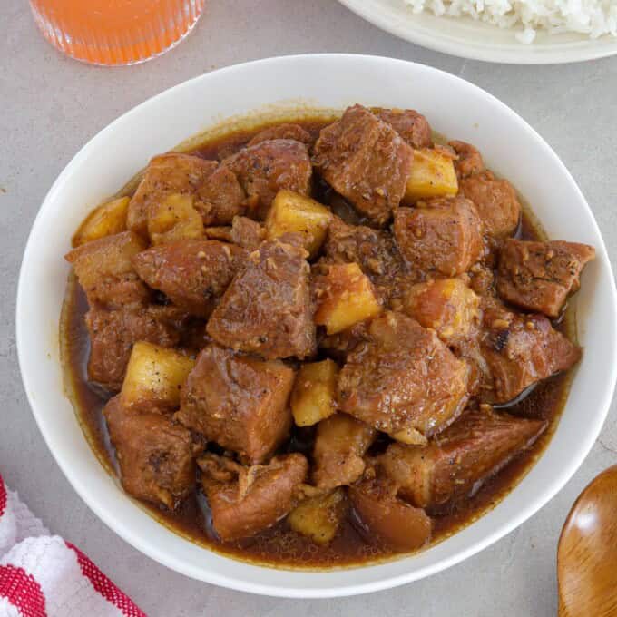 pork adobo with pineapple in a white serving bowl with a plate of steamed rice in the backround.
